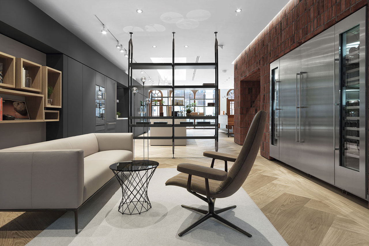High-End-Bildbearbeitung – Visual: Gaggenau Showroom, eleganter offener Raum mit Einbaukühlschränken aus Edelstahl in Ziegelwand, Ledersofa, Lounge Chair, Glasbeistelltisch, Regal-Raumtrenner, Holzboden