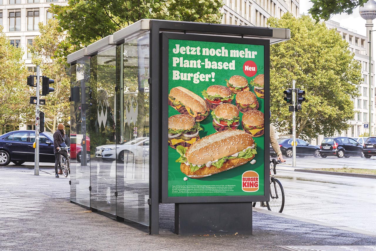 Zu sehen ist ein beleuchtetes, grünes City-Light an einer Bushaltestelle in einer freundlichen Szene einer Innenstadt mit Bäumen und hellen Gebäuden. Das City-Light-Motiv besteht diversen plantbased (vegetarischen) Burgern sowie dem Logo der Marke Burger King.